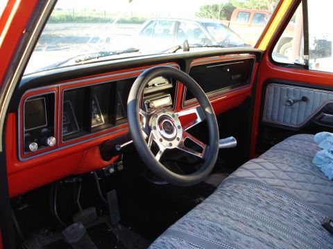 1979 Ford F-150 - Interior of an orange-red classic Ford pickup shot from the open driver's door, showing the body-color dash, black instrument cluster bezel…
