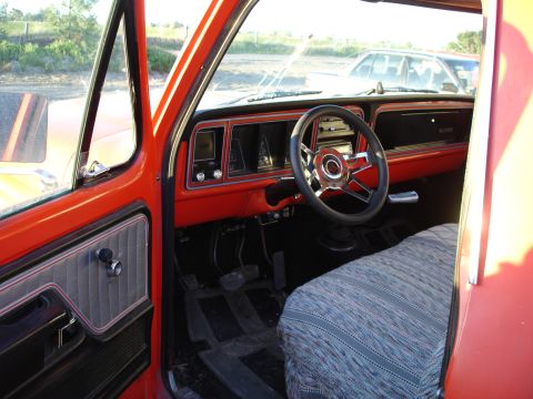 1979 Ford F-150 - Interior of an orange-red classic Ford pickup shot through the open driver's door, showing the body-color dash and door panel with gray tuc…