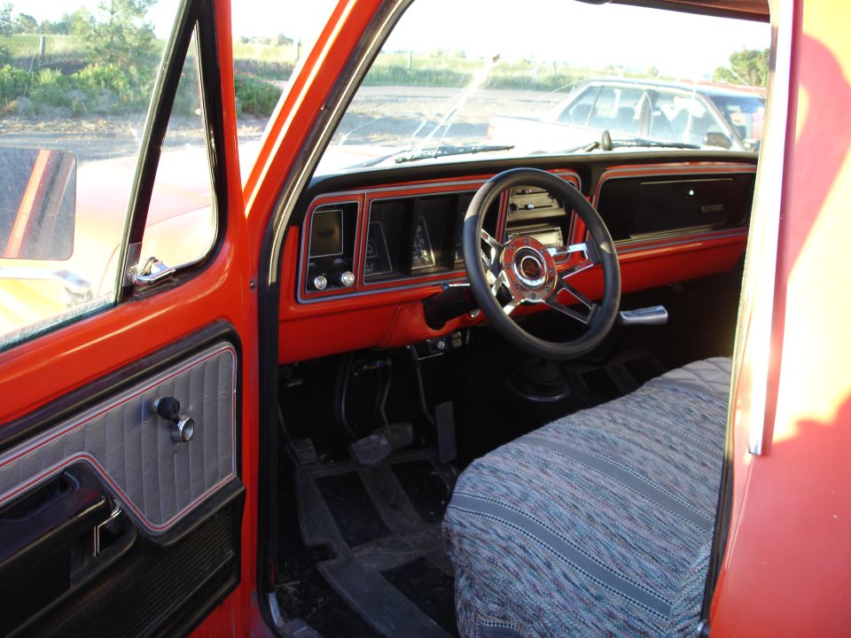 Driver's-side interior of orange-red classic Ford pickup with chrome steering wheel and gray bench seat