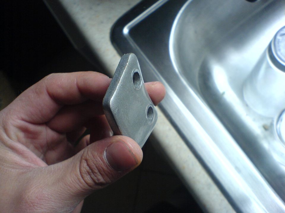 Hand holding a small, diamond-shaped metal part with two holes near a sink