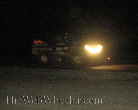 1990 Ford Bronco - off-road race truck numbered 300 sits in the dark desert at night with bright headlight glare and a red taillight visible.