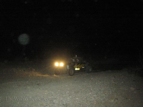 1990 Ford Bronco - off-road buggy with two bright headlights sits on a dirt road in near-total darkness at night.
