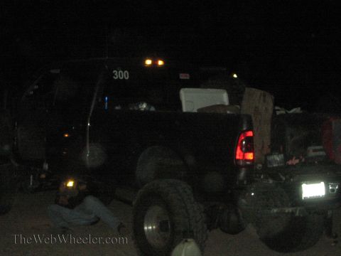 1990 Ford Bronco - dark vehicle numbered 300 is parked at night with a spare tire and tools nearby and a person lying underneath it during a repair.