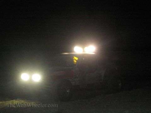 1990 Ford Bronco - off-road race truck runs at night with its roof-mounted light bar and headlights blazing across the dark desert.