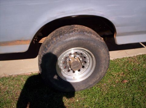 1995 Ford Bronco - Rear wheel and quarter panel of a white truck parked on grass, with a worn all-terrain tire on a polished aluminum slotted wheel and a manu…