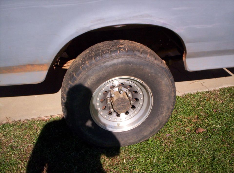 Worn all-terrain tire on aluminum wheel under white truck's rusty rear fender