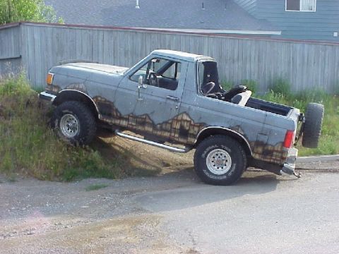 1989 Ford Bronco - Passenger-side rear three-quarter view of a lifted gray full-size Ford Bronco with its hardtop removed, climbing a grassy embankment beside…