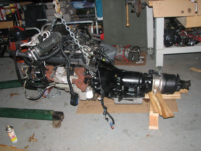 Engine and transmission assembly suspended by chains, resting on wooden blocks and a green stand