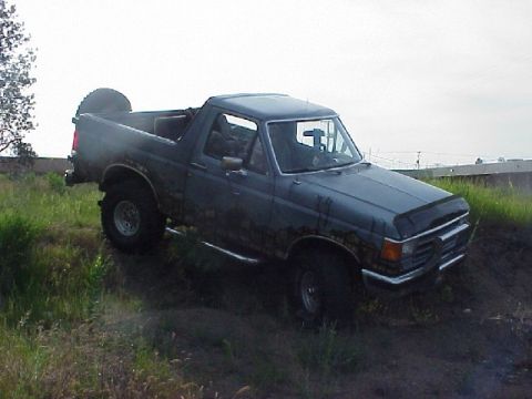 1989 Ford Bronco - Passenger-side front three-quarter view of a lifted gray full-size Ford Bronco with the hardtop removed, driving through tall grass and dir…