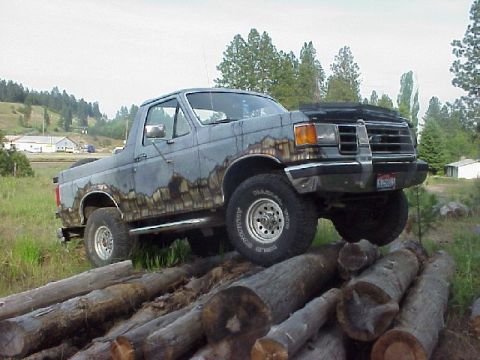 1989 Ford Bronco - This heavily rusted Ford Bronco is positioned on a stack of logs, with a rural landscape in the background.