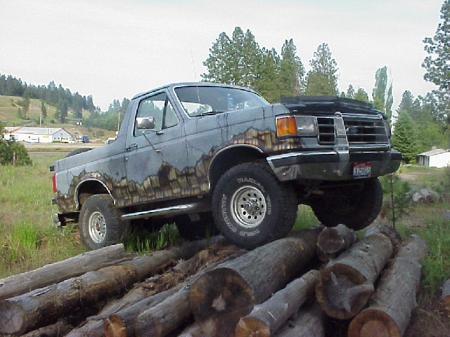 Gray Ford Bronco with significant rust on the lower body panels, angled on a pile of logs
