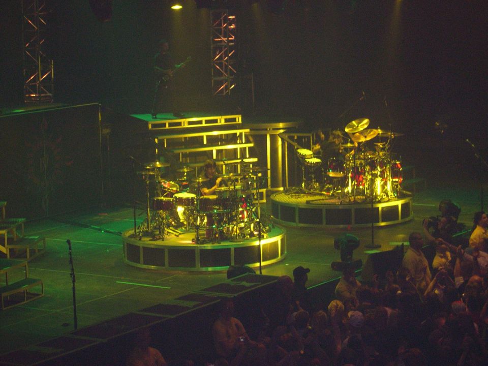 Two drummers play on elevated circular stages with multiple drum kits and cymbals under stage lights