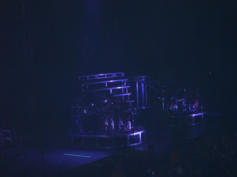 Stage setup with two drum kits on circular platforms under blue lighting. SUPERMOTORS.NET logo in lower right