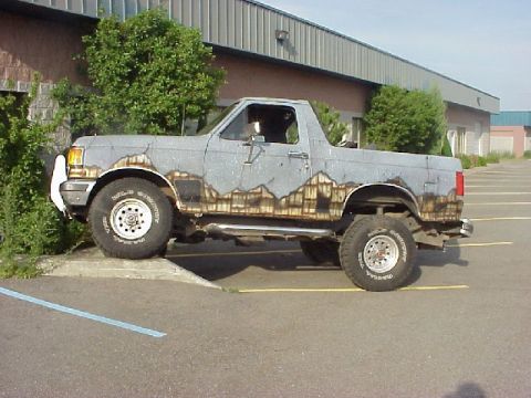 1989 Ford Bronco - Passenger-side profile of a lifted full-size Ford SUV with the top and doors removed, flexing with the front wheel up on a parking-lot curb…