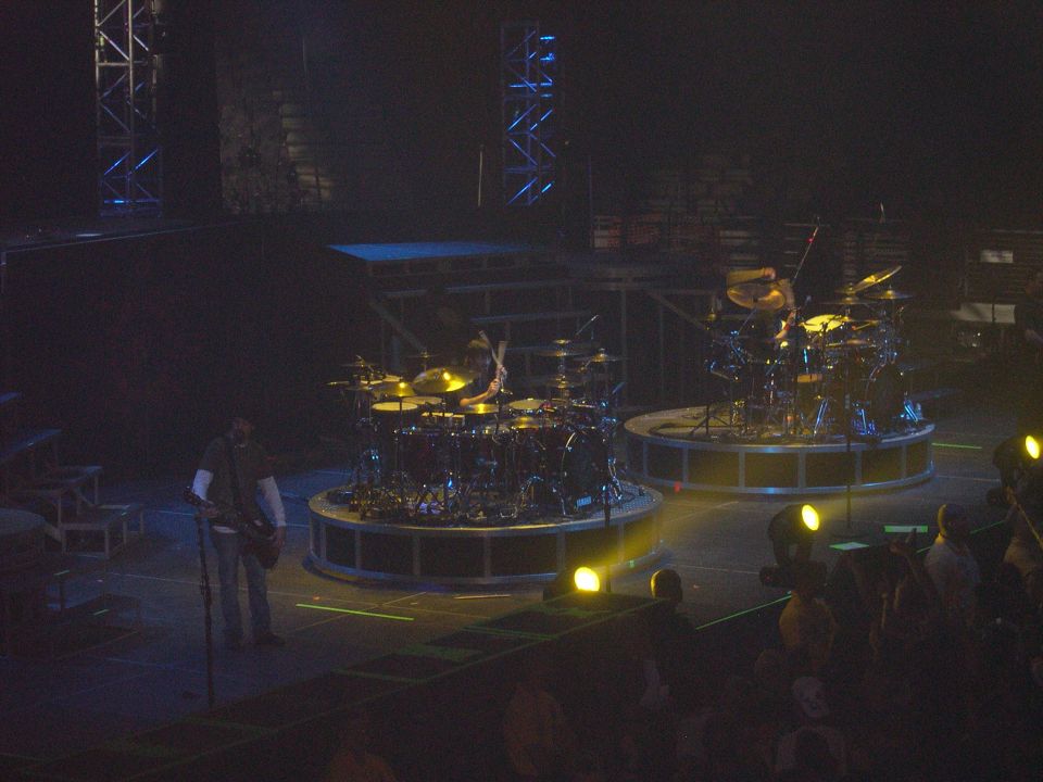 Concert stage with two drum kits, musicians, and audience members visible