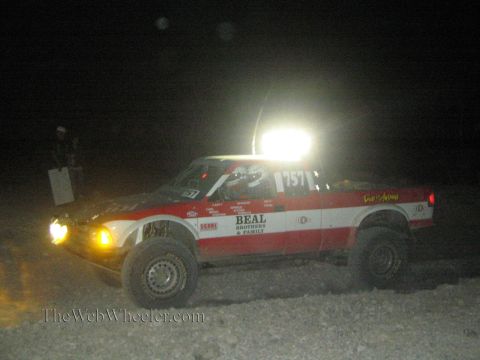 1990 Ford Bronco - red and white off-road race truck numbered 1571 with Beal Brothers decals and a bright roof light bar runs at night.