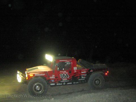 1990 Ford Bronco - red Hummer-style off-road race truck numbered 864 with BFGoodrich and Baja decals runs through the desert at night.