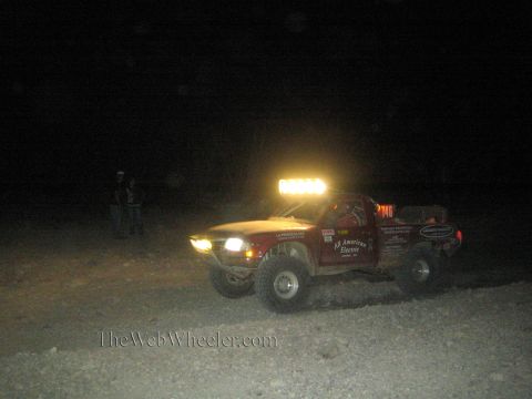 1990 Ford Bronco - red off-road race truck with an All American Electric livery and bright roof light bar runs at night with spectators nearby.