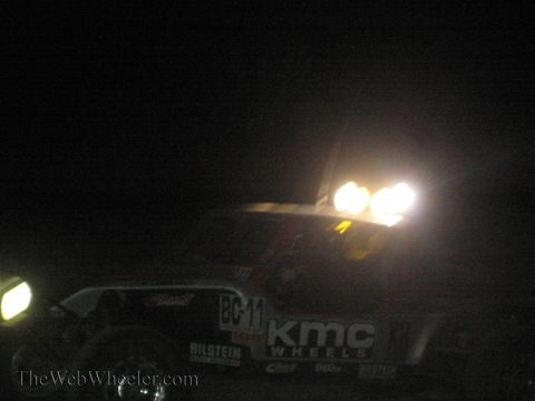1990 Ford Bronco - off-road race vehicle showing KMC Wheels and Bilstein decals shines its lights into the camera in the dark desert.