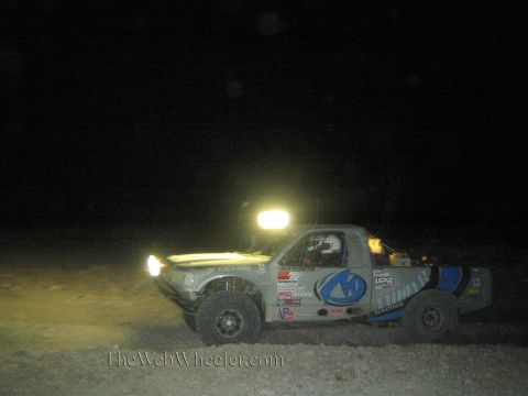 1990 Ford Bronco - off-road race truck with a blue logo on the door and a bright roof light bar runs through the dark desert at night.