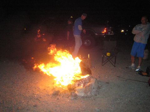 1990 Ford Bronco - Several people stand around a large blazing campfire at a desert camp at night, with a vehicle and folding chair nearby.