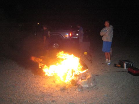 1990 Ford Bronco - man with crossed arms stands beside a blazing campfire near a pickup truck at a desert camp at night.