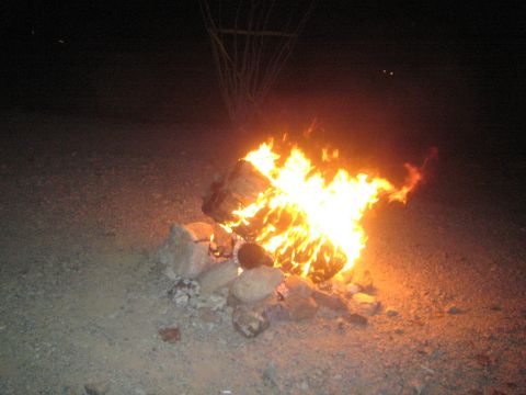 1990 Ford Bronco - large campfire ringed with rocks burns brightly on the gravel ground of a dark desert camp at night.
