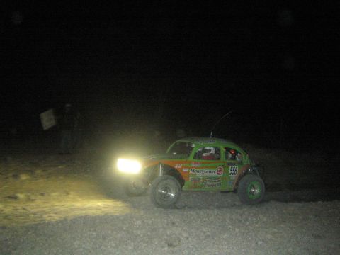 1990 Ford Bronco - green Volkswagen Beetle-based Baja race car numbered 555 with MoneyGram decals runs at night with a spectator nearby.