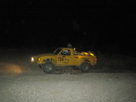 1990 Ford Bronco - yellow off-road race truck numbered 724 kicks up dust as it races across the dark desert at night.