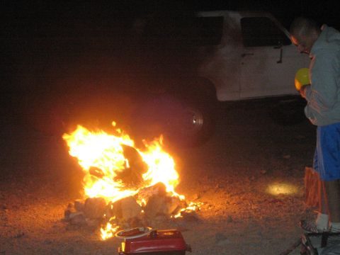 1990 Ford Bronco - campfire burns on the desert ground beside a white truck while a person in a hoodie stands at right at night.