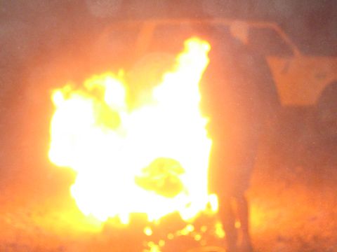 1990 Ford Bronco - Bright orange flames from a campfire fill the frame with the faint silhouette of a vehicle behind at night.