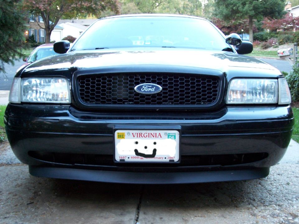 Front view of a black 2001 Ford Crown Victoria. It has a black honeycomb grille with a Ford logo and a Virginia license plate with a smiley face drawn on it