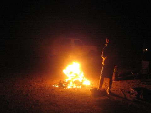 1990 Ford Bronco - man stands beside a glowing campfire near a parked truck at a desert camp at night.