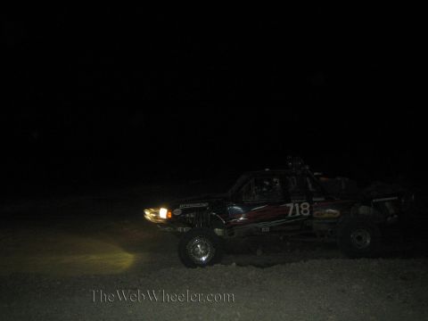 1990 Ford Bronco - dark off-road race truck marked 718 races at night with one headlight illuminating the desert ground.