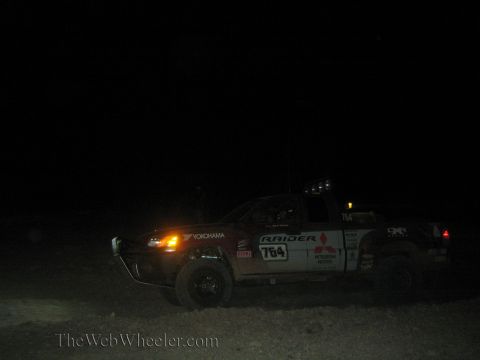 1990 Ford Bronco - white Mitsubishi Raider off-road race truck numbered 764 with light bar runs through the dark desert at night.
