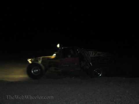 1990 Ford Bronco - off-road race truck marked 717 speeds across dark desert terrain at night, lit only by its headlights.