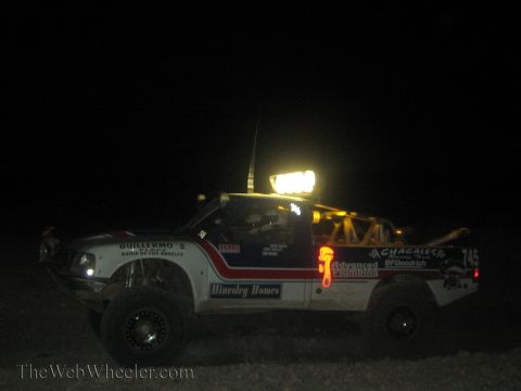 1990 Ford Bronco - white sponsor-covered off-road race truck numbered 745 with a glowing roof light bar sits in the dark desert at night.