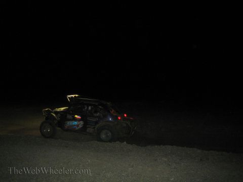 1990 Ford Bronco - off-road race buggy with a roof light bar drives across dark desert terrain at night.