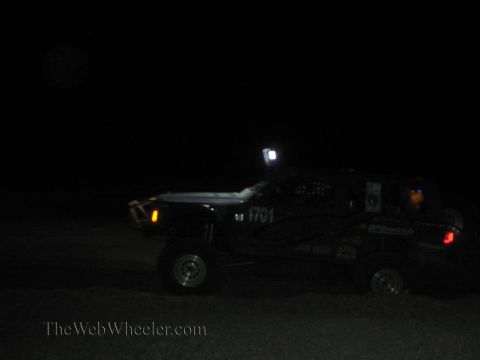 1990 Ford Bronco - dark off-road race truck marked 1701 with a bright roof light races across the desert at night.