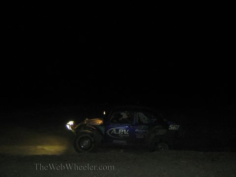 1990 Ford Bronco - blue Baja Bug race car numbered 561 with sponsor decals drives through the dark desert at night.