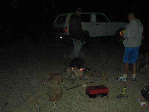 1990 Ford Bronco - Two men stand near a small campfire and a portable grill beside a white SUV at a dim desert camp at night.