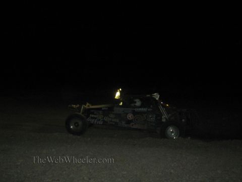 1990 Ford Bronco - off-road race buggy covered in beverage sponsor decals speeds across dark desert terrain at night.