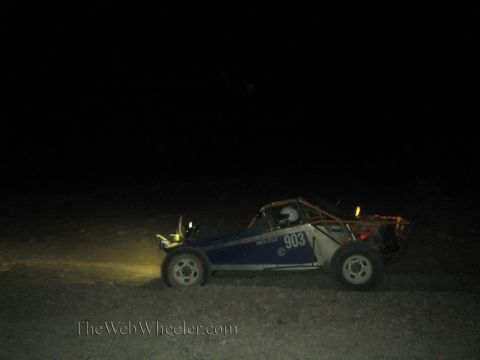 1990 Ford Bronco - blue tube-frame off-road race buggy numbered 903 drives across the dark desert lit by its headlights at night.