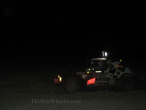 1990 Ford Bronco - off-road race car numbered 1100 with checkered livery and a roof light runs in the dark desert at night.
