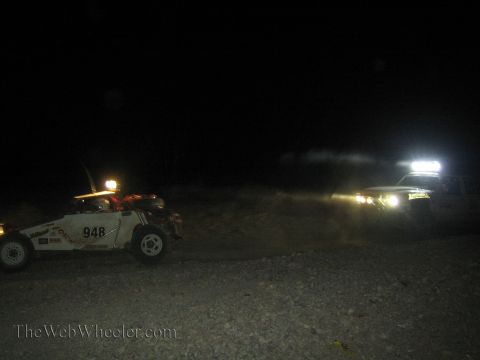 1990 Ford Bronco - off-road race car numbered 948 sits at left as another vehicle with bright lights approaches through dust at night.