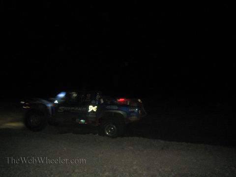 1990 Ford Bronco - blue off-road race truck with a Shepard decal and a red light drives across dark desert terrain at night.