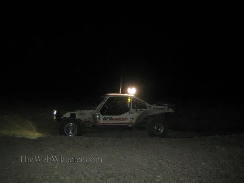 1990 Ford Bronco - white off-road race car with BFGoodrich decals and a roof light runs across the dark desert at night.