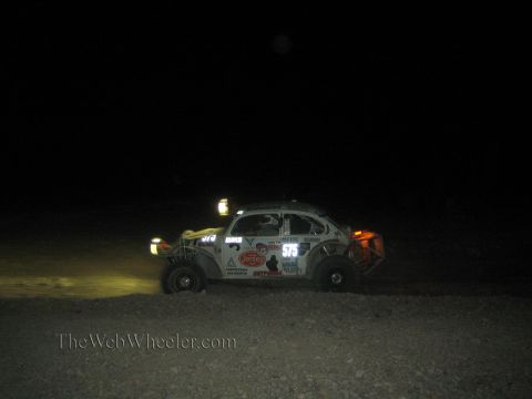 1990 Ford Bronco - white Volkswagen Beetle-based Baja Bug race car numbered 575 runs through the dark desert at night.