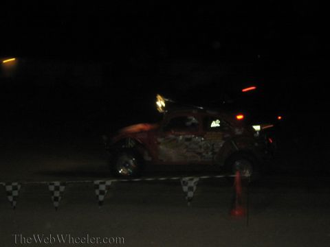 1990 Ford Bronco - red Baja Bug race car with stickers passes a checkered course barrier and traffic cone at night.