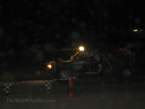 1990 Ford Bronco - off-road race buggy with a roof light passes a checkered course barrier and cone amid dust at night.